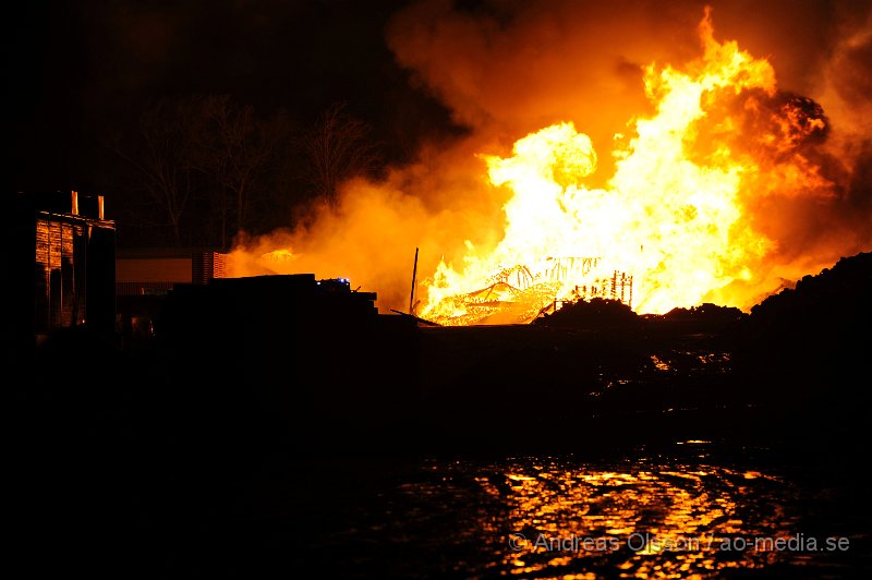 _DSC8505.JPG - Vid 21.40 larmades räddningstjänst, ambulans och polis till Gyllsjö trä i Stidsvig där man hade en fullt utvecklad brand i ett pall lager med ungefär 10 000 trä pallar. Lagret var helt övertänt när räddningstjänsten kom fram till platsen och man inrikta sig direkt på att skydda omkring liggande byggnader. Bland annat produktionslokalen som ligger ungefär 20 meter från pall lagret. Den kraftiga strålnings värmen från branden gjorde att det började brinna lite i fasaden på produktionslokalen men man lyckades begränsa branden och skydda byggnaden. Vid tre tiden på natten fick man en brand uppe i taket på produktionslokalen och man fick sätta in fler resurser, men kallade in en hävare och skickade upp folk på taket för att begränsa branden och kontrollera så den inte kunde sprida sig. Det arbetet gav också snabbt resultat och skadorna blev bregränsade. Ingen person har kommit till skada men lager byggnaden är helt nedbränd och produktionslokalen har fått skador i ena fasaden. Räddningstjänsten hade flera stationer på plats under kvällen/natten, Klippan, Åstorp, Ljungbyhed; Örkelljunga, Ängelholm, Helsingborg.